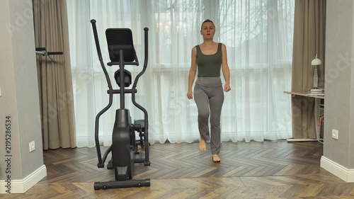 Woman in sports top and leggings walks calmly in place with arms lifted overhead. Light recovery movement in living room after cardio workout.