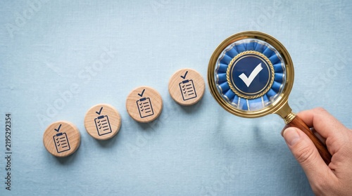 A hand holding a magnifying glass over a checklist on a wooden block, symbolizing quality control and verification. The blocks are arranged in a line, suggesting progress and completion.
