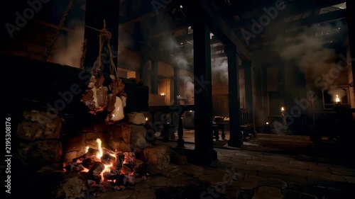 Dimly lit interior of medieval tavern with meat roasting over stone fireplace hearth and smoky air, wide shot