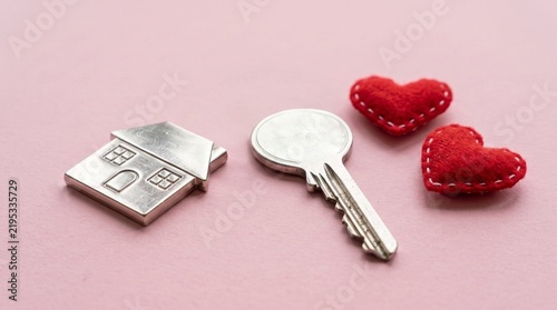 Metallic house shaped keychain placed beside shiny silver door key and two small soft red felt heart ornaments isolated on a light pink textured background
