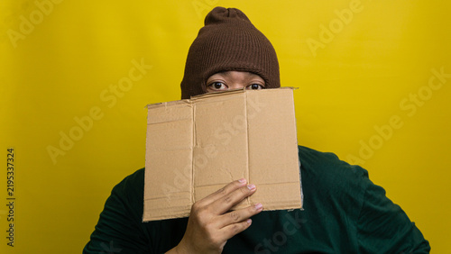 Fotografía Young Asian male wearing brown balaclava obscures lower visage with blank corrugated board against vivid yellow backdrop
