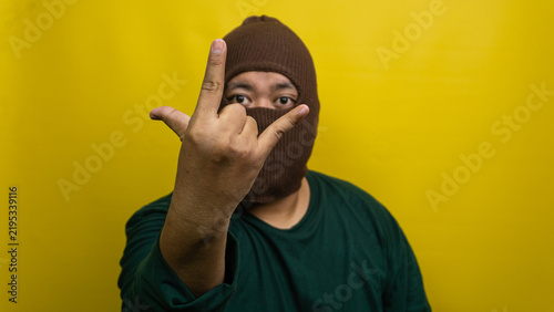 Bild auf Leinwand Young Asian male wearing brown balaclava displays rock and roll hand gesture against vivid yellow backdrop