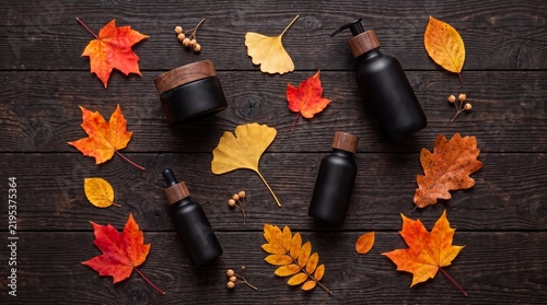 Matte black cosmetic bottles and diverse colorful fall leaves scattered on dark wooden board, seasonal premium organic skincare, rustic autumn composition