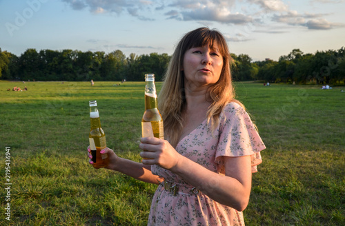 Obraz na plátně Young woman holding two beer bottles while standing in a sunny grassy park at go