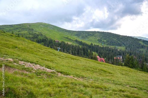 Vast green mountain slopes feature scattered evergreen trees, ski lift towers, and small red-roofed buildings under a cloudy overcast sky. Dragobrat, Carpathians, Ukraine