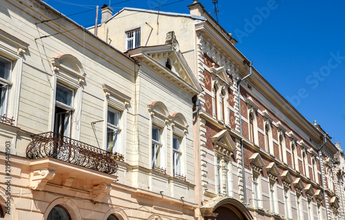 Elegant historic street-facing building facades feature ornate moldings, a wrought iron balcony, arched windows, and decorative red brick accents under a clear blue sky. Chernivtsi, Ukraine