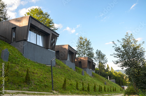 Modern dark wood clad cabins sit in a row along a grassy slope, framed by surrounding green trees and a clear blue sky with light clouds above