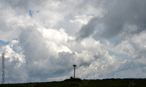 Solitary wooden directional signpost stands silhouetted atop a grassy hilltop beneath a vast, dramatic overcast sky filled with heavy grey and white clouds
