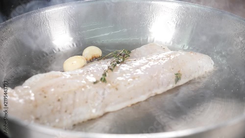 White fish fillet frying in stainless steel pan with garlic cloves and fresh thyme. Rising steam, professional kitchen cooking process, delicious seafood meal preparation Slow motion 4k video