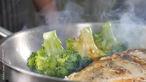 Close-up of chef using gas torch flame to char broccoli in stainless steel pan with fish fillet. Rising steam, professional kitchen cooking process, healthy meal preparation Slow motion 4k video
