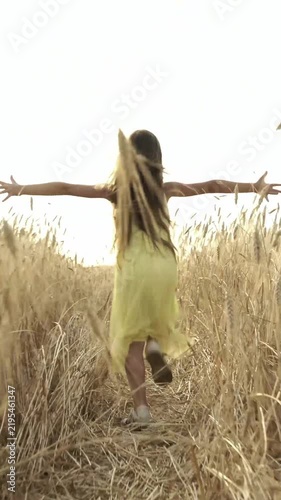 Young girl in yellow dress runs freely through golden wheat field at sunset, arms wide.