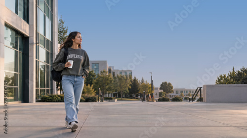 A female university student walks across a spacious campus plaza carrying a backpack and takeaway cup, with modern glass buildings and trees beneath a clear sky.