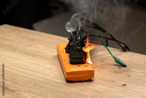 Overloaded orange power strip burning with fire and smoke with green jumper clip wire on wooden table, dangerous short circuit fire hazard concept.
