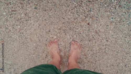 Male feet standing on clear water and sandy beach, top-down view. 4K