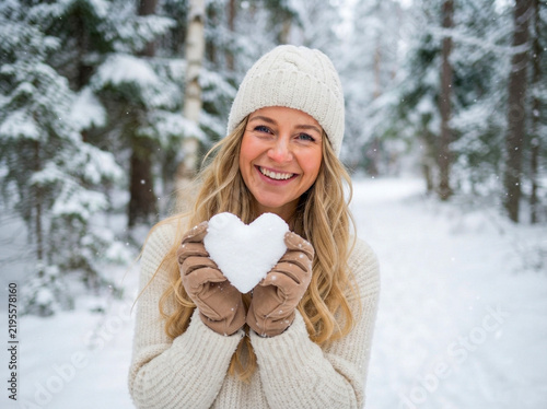 Smiling woman holding a heart shaped snow snowball in a snowy winter forest wearing a white knitted hat and sweater during a cold seasonal day