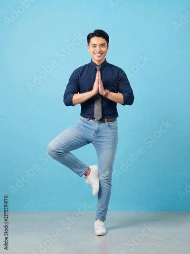 Businessman practicing yoga pose wearing formal shirt and tie while standing on one leg isolated against a solid light blue background in a professional studio