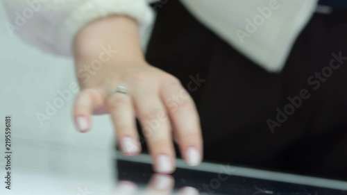 Close-up of a woman's hand touching a tablet. Fingers have light pink nails. Ring visible on her finger. Blurred background suggests office setting. Focus on hand and device interaction