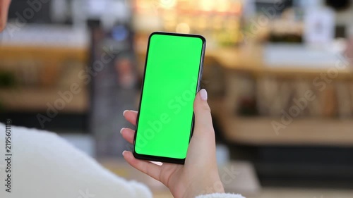 Person holds smartphone with green screen. Background shows blurred cafe interior. Hand grips device steadily. Scene focuses on phone display only