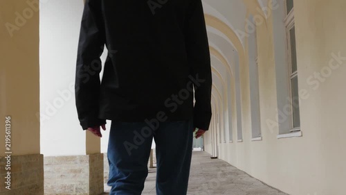 Adult man in black jacket jogs away down symmetrical arched passageway with columns, camera follows from behind in tracking motion, architectural perspective.