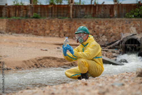 Fotografie Environmental worker in protective suit collects water directly from polluted stream near industrial drainage site for contamination testing, water quality analysis, pollution research