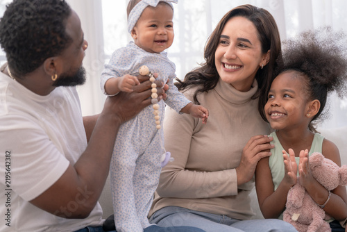 A fun-loving father with his baby holds up his giggling baby to play on a sunlit white sofa, while the daughter cheers happily in a warm welcoming home ideal for parenting and wellbeing.
