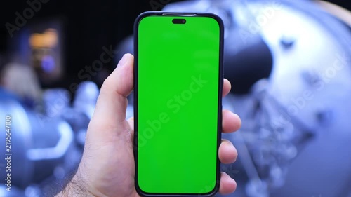 Hand holds phone with green screen. Background shows blurred futuristic equipment. Hand grips device firmly. Focus remains on phone display