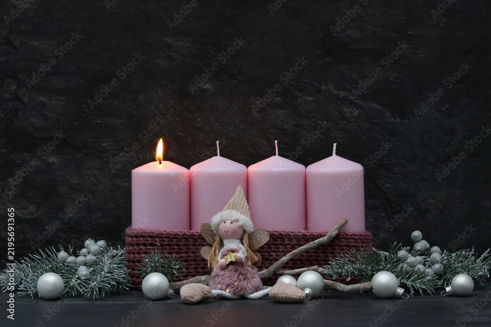 Fototapeta premium First Sunday of Advent: A burning pink candle on a modern Advent wreath featuring an angel figure, fir branches, and silver baubles against a dark background, with copy space.
