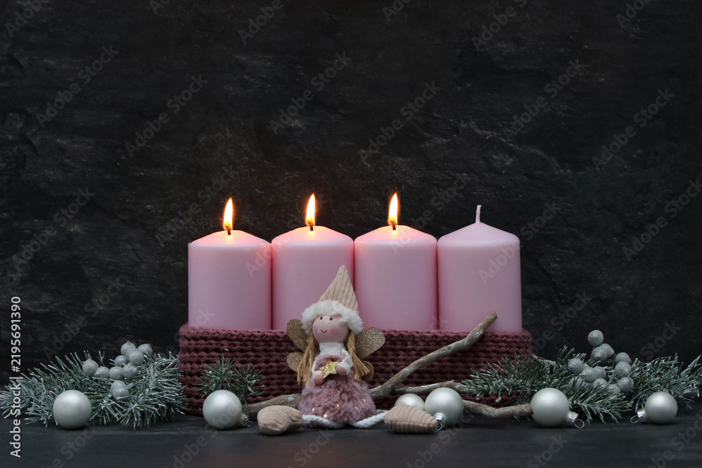 Fototapeta premium Third Sunday of Advent: Three burning pink candles on a modern Advent wreath featuring an angel figure, fir branches, and silver baubles against a dark background, with copy space.