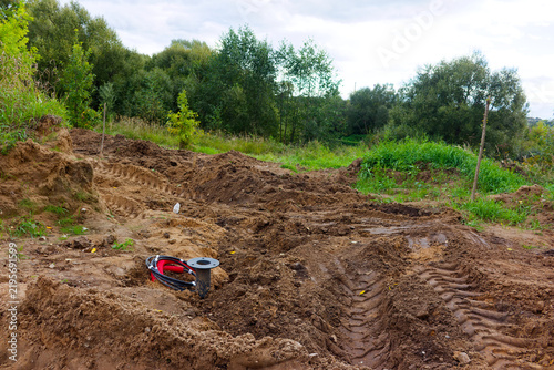 Industrial black cable with red sleeve buried into rough brown soil at active construction site with heavy machinery tire tracks amidst a green natural landscape background