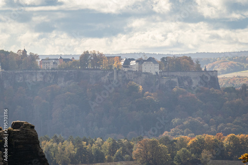 Fotografie Königstein Fortress in Saxon Switzerland, Germany, perched on a flat-topped sandstone mountain above an autumn forest valley under a cloudy sky