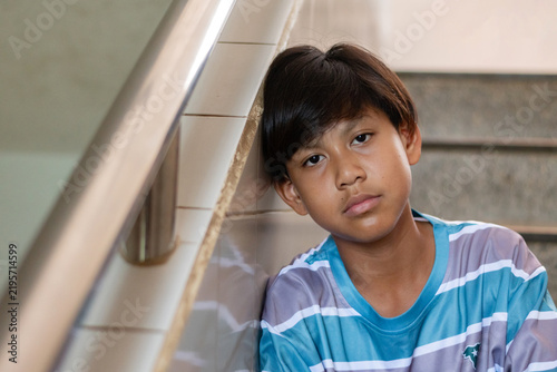 A tired teenage boy in a striped shirt leans his head against a tiled wall while sitting on stairs.
