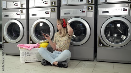 A young woman with blonde hair is seated on the floor of the laundry room, near a basket of garments. She is listening to music on her mobile device as she waits for her clothes to be cleaned.
