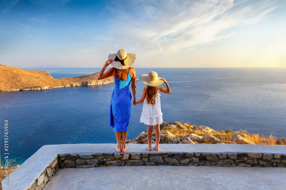 Obraz premium Summer family travel concept with a mother and her daughter in dresses enjoying the sunset view over the ocean from their terrace, Cyclades, Greece