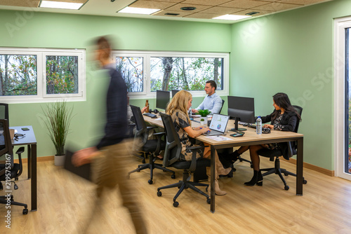 Coworkers collaborating and working at a long desk in a bright, modern office space with green walls, a light wooden floor, and a blurred person moving through the foreground