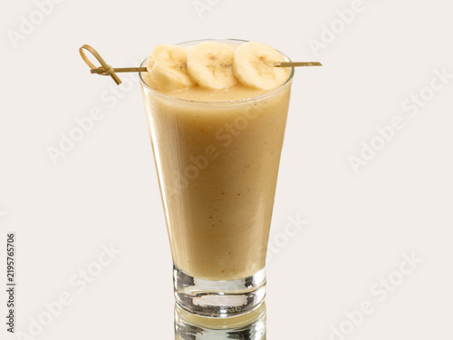 Tall glass of creamy banana smoothie topped with fresh banana slices on a skewer, shot on a light background.