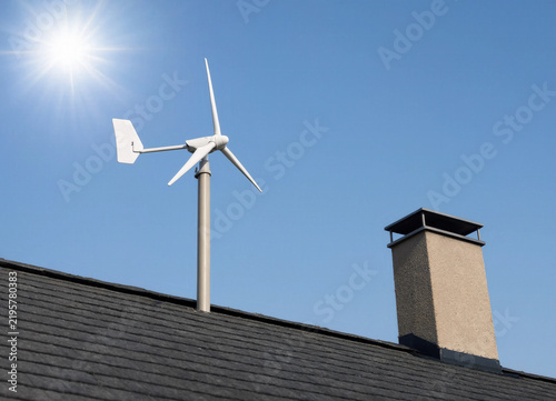 A small white domestic wind turbine, installed on the dark tiled roof next to a chimney, under the bright sun.