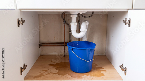Water leaking from a pipe under the sink with a blue bucket catching the water
