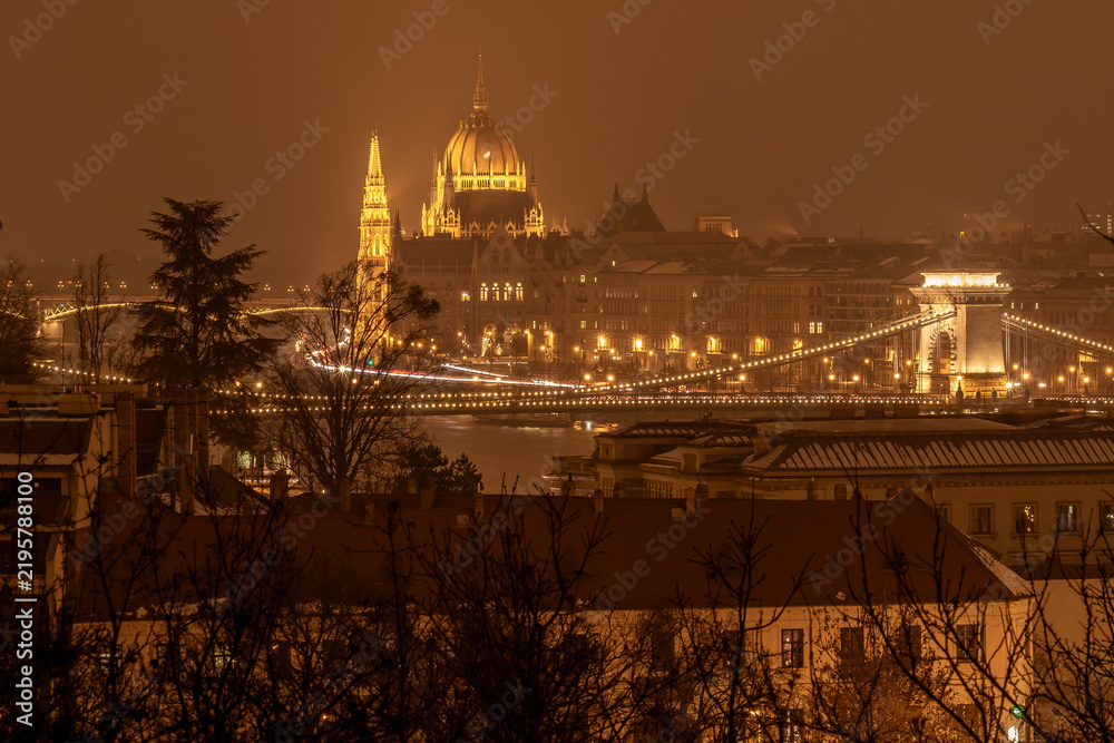 Obraz premium Illuminated Hungarian Parliament and Szechenyi Chain Bridge glowing golden against an atmospheric, misty night sky in Budapest.