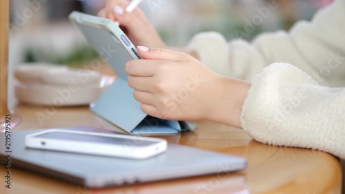 Person uses stylus on tablet at wooden table. Phone and laptop sit beside tablet. Hands wear white knitted sweater. Warm indoor lighting. Blurred background with natural tones