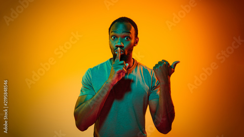 Konstfotografi Secretive man makes silence gesture and points aside in orange studio with alert expression
