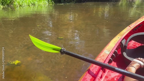 POV kayaking on a shallow calm river, red kayak with paddle gliding through clear water surrounded by green summer nature, outdoor recreation footage in 4K.