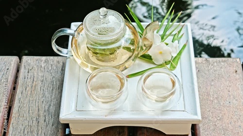 Glass teapot with herbal tea and two cups on white tray, wooden deck by koi pond with koi carp, spa wellness relaxation concept, outdoor tropical garden setting, nobody. Slow motion.