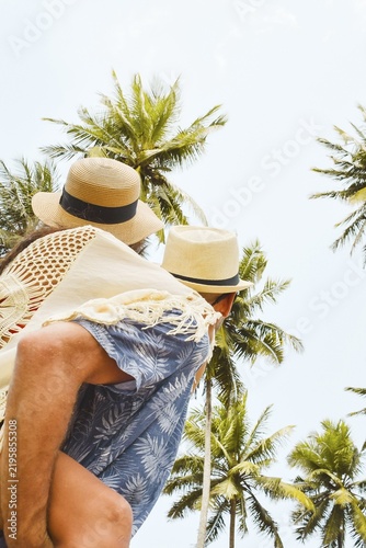 Close up young man giving woman piggyback ride laughing together looking up under towering coconut palm trees in tropical island beach resort destination sunny summer day. Ad promotional banner