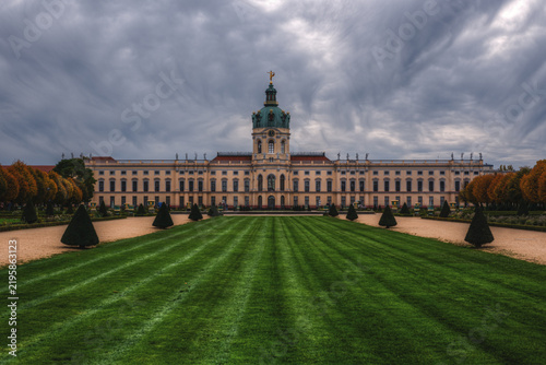 View of Charlottenburg Palace with its manicured green lawn, symmetrical topiary trees, and historic baroque architecture under a dramatic cloudy sky in Berlin, Berlin, Germany.