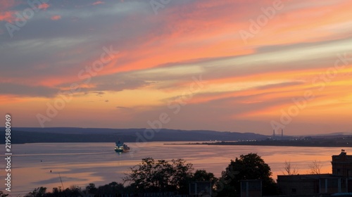 Obraz na płótnie Glowing orange and pink sunset clouds stretch above calm coastal water with a small boat and dark foreground silhouettes