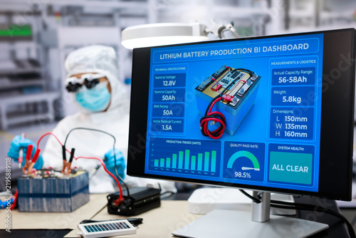 Research worker in protective gear assembles a battery beside a monitor lithium battery production data, quality testing, capacity and safety status in a modern laboratory.