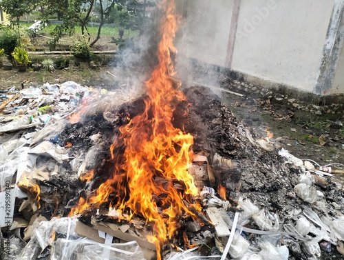large pile of mixed waste is burning outdoors, with tall orange flames and thick gray smoke rising into the air