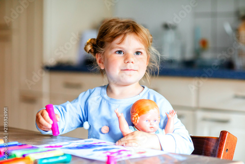 Little girl drawing with colorful markers while playing with a doll at home, enjoying a creative indoor activity and early childhood learning