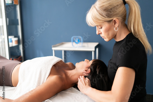 Female therapist performing neck and shoulder massage on relaxed adult woman in treatment room with soft natural light