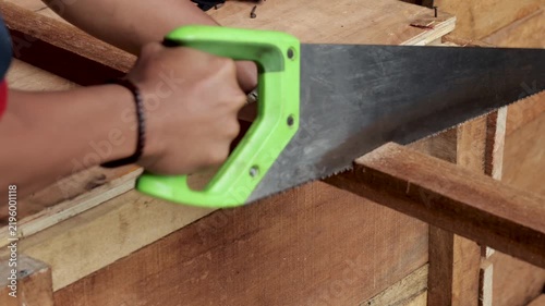 Carpenter cutting wooden plank using manual hand saw inside carpentry workshop.